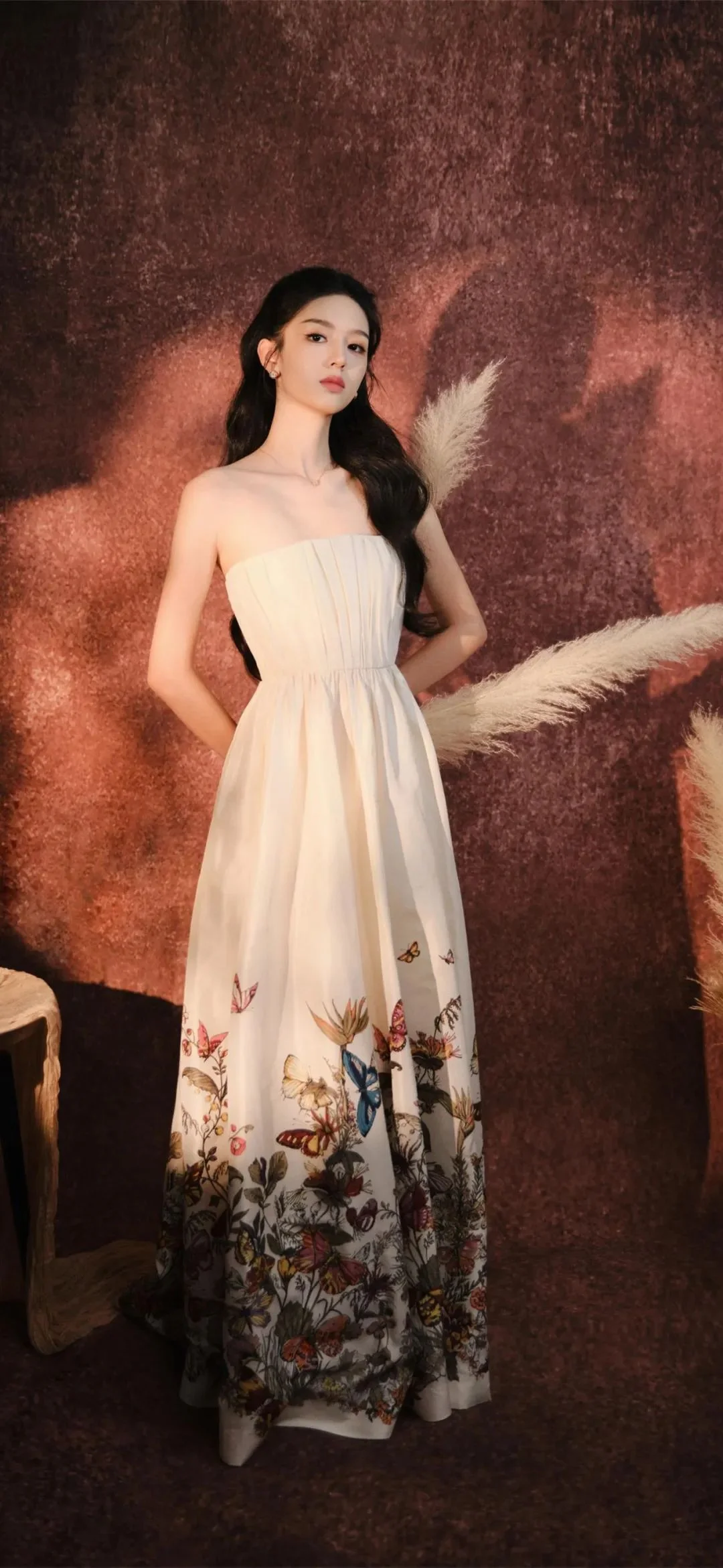 一名成年女性身穿带有花卉图案的长裙，站在棕红色背景和羽毛装饰前。