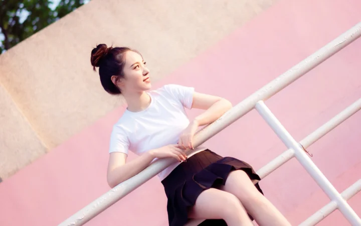 一名成年女性穿着白色上衣和黑色裙子，靠在白色栏杆旁，背景呈粉色。