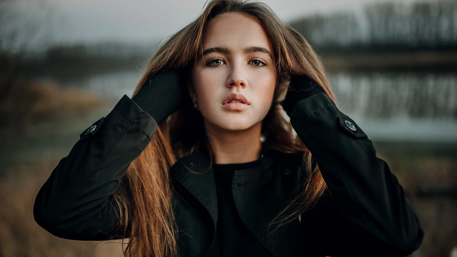 一名长发女性身穿黑色外套，在户外湖边背景前直视镜头。
