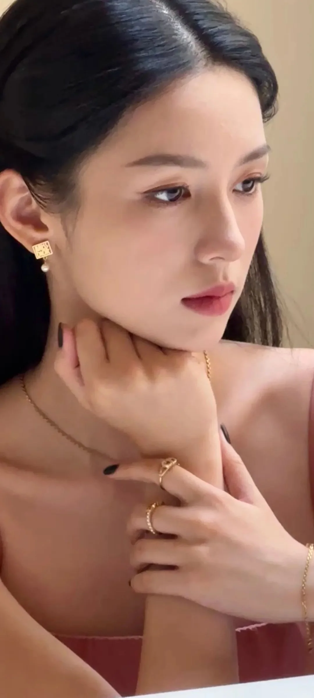 一名女性在柔和室内光线下侧脸凝视，手托着颈部并佩戴金色耳饰、项链和戒指。