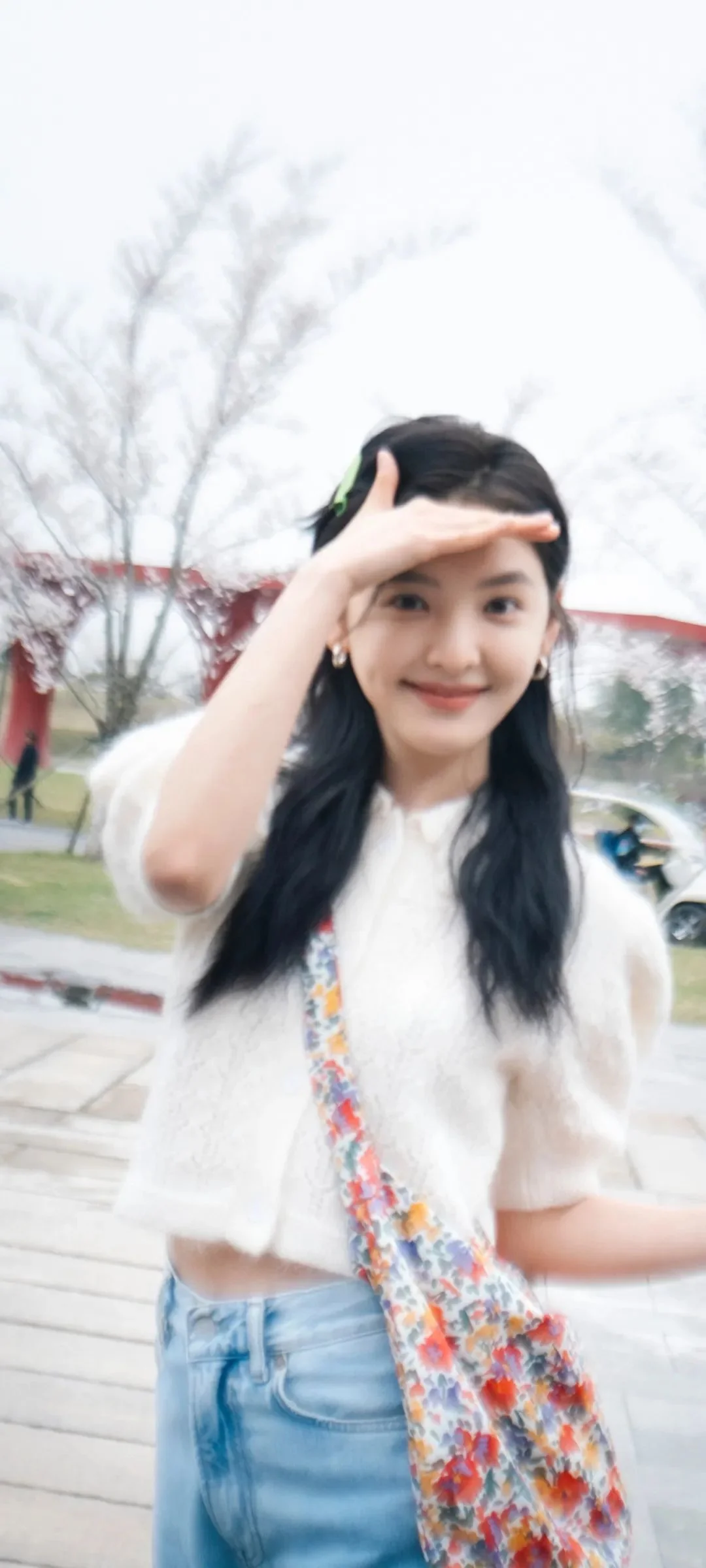 긴 머리의 여성이 야외에서 미소 지으며 한 손을 들어 햇빛을 가리고 있다.