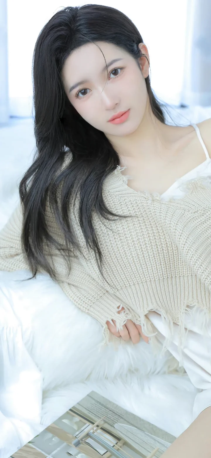 一名长发女性穿着白色针织衫坐在明亮的室内环境中。