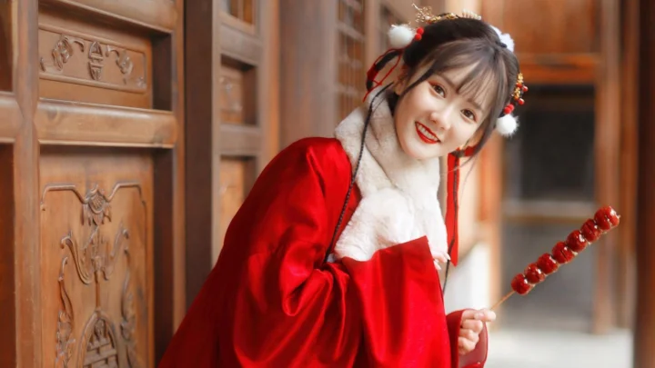 一名身穿红色传统服饰的女性站在木质门前，手持红色糖葫芦并面带微笑。