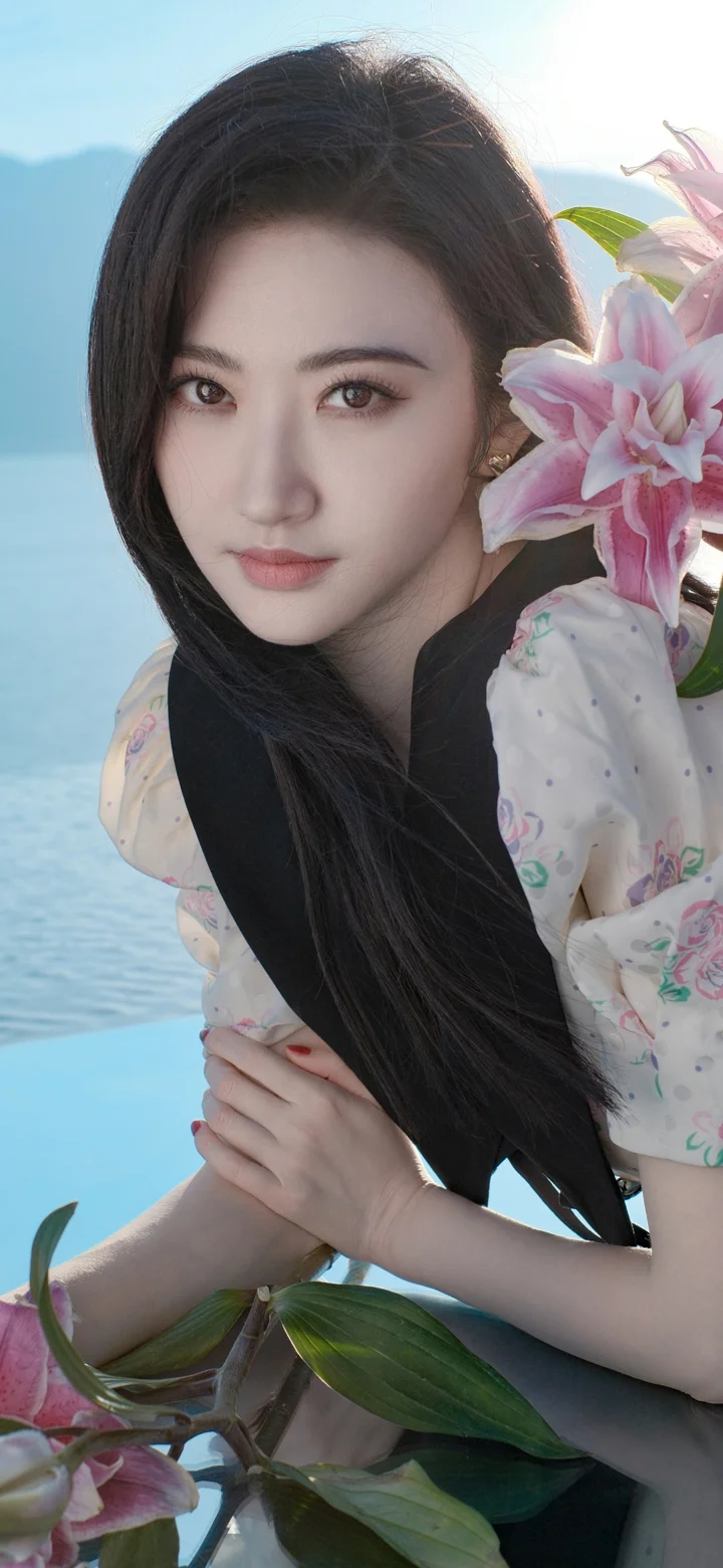 一名长发女性在水边手捧粉色花朵，背景映着蓝色水面和远山。