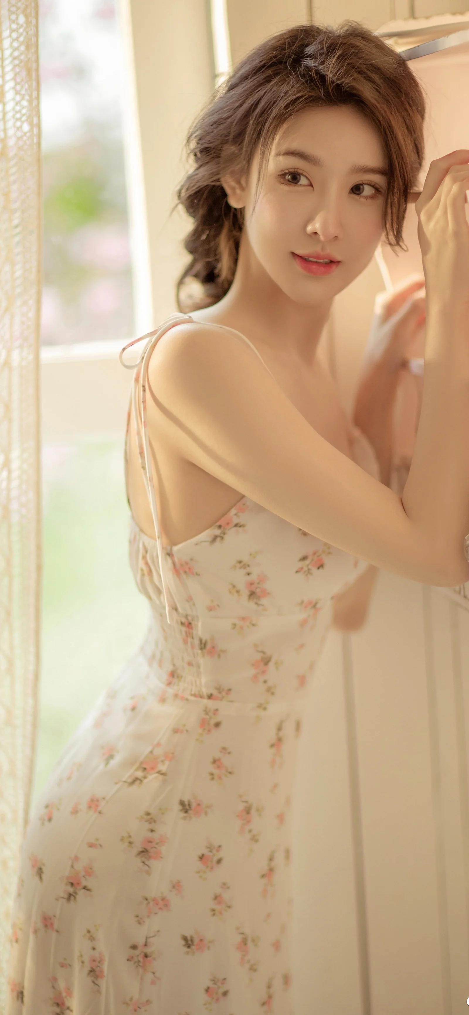 一名成年女性穿着碎花连衣裙站在明亮窗边回望镜头。