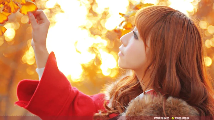 一名身穿红色外套的女子在温暖的秋日阳光中伸手触碰橙色树叶。