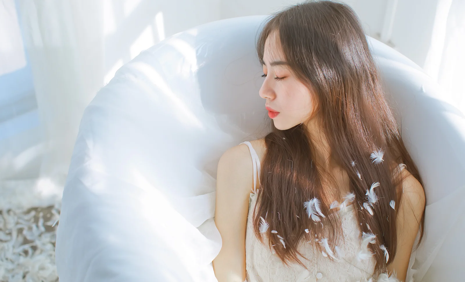 긴 머리의 여성이 눈을 감고 흰색 의자에 앉아 있으며 머리카락과 옷에 작은 흰 깃털이 보인다.