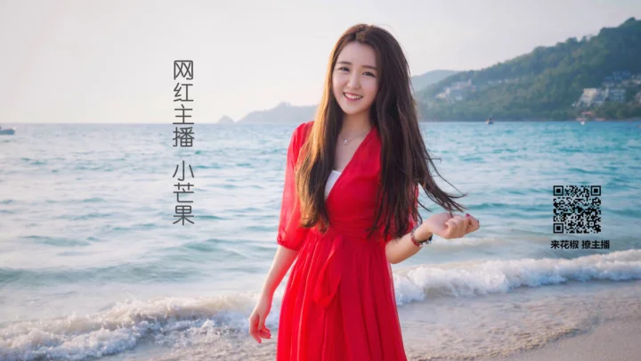 一名身穿红裙的成年女子站在海滩上，身后是海水、山坡和远处的建筑。