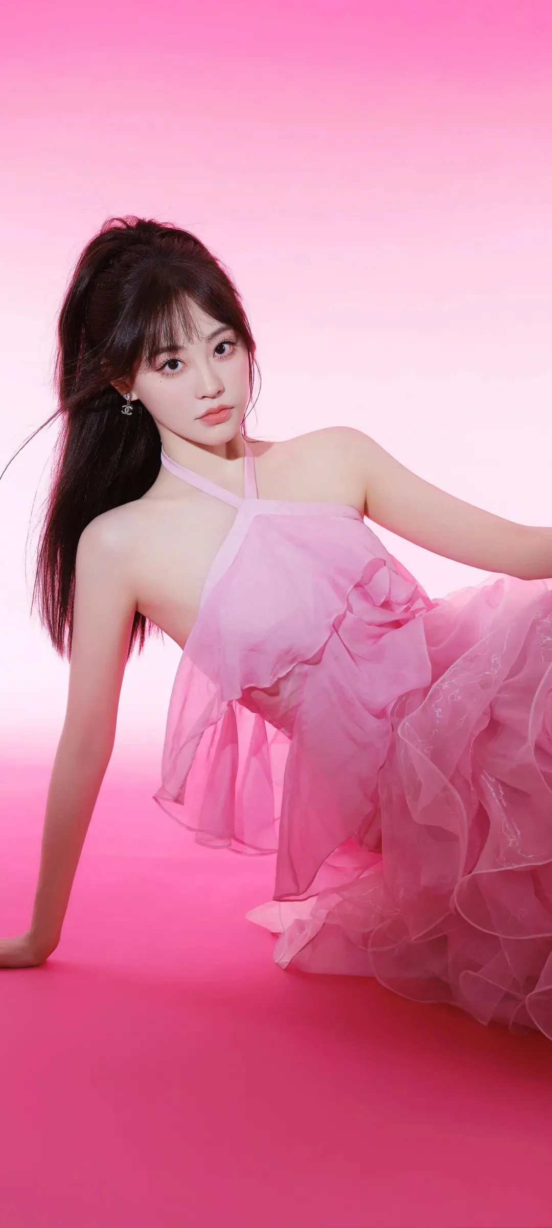 一位成年女性身穿粉色连衣裙，在粉色背景前侧身坐姿面向镜头。