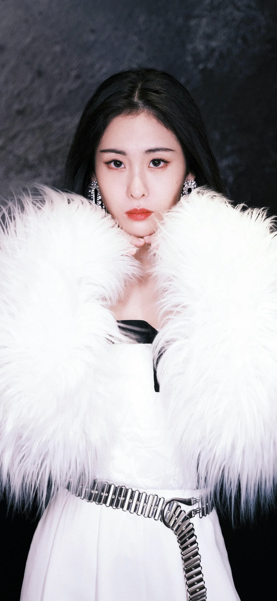 一名黑发女性身穿白色蓬松外套，站在深色背景前直视镜头。
