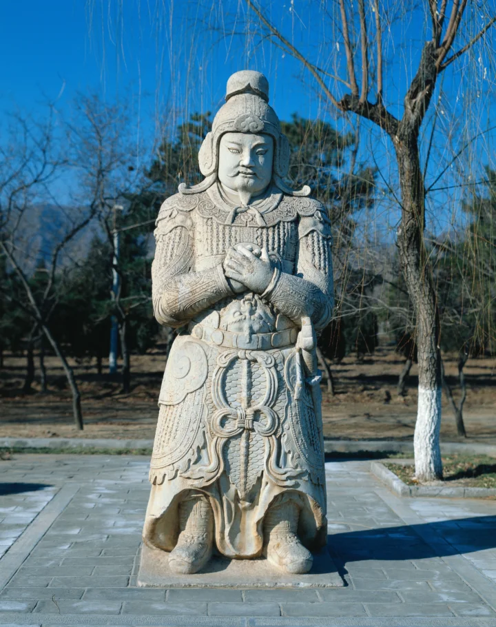 一尊雕刻精细的古代武将石像矗立在蓝天下的户外石板路上，周围环绕着树木。