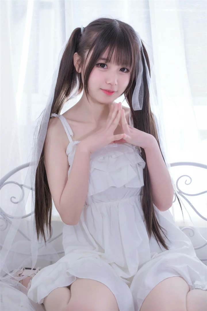 긴 머리의 여성이 흰색 원피스를 입고 밝은 커튼 앞에 앉아 가슴 앞에서 두 손을 모으고 있습니다.