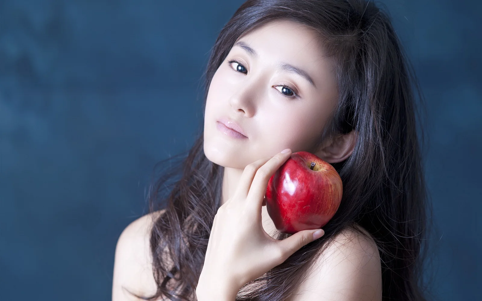 一名成年女性手持红苹果靠近脸颊，在蓝色背景前摆姿势。