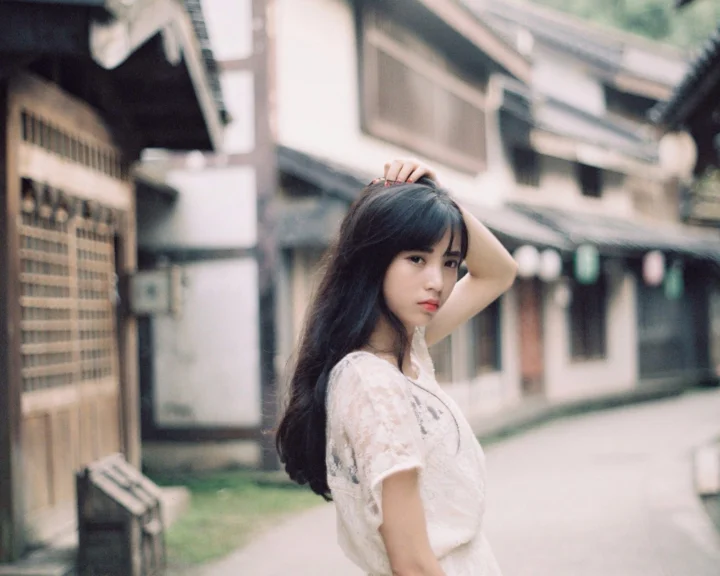 一名长发女子站在老街巷中，抬手触碰头发，背景是虚化的建筑与街道。