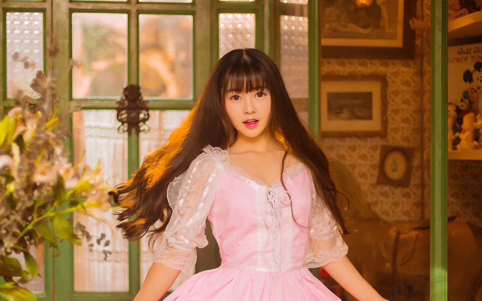 長い髪の女性がピンクのドレスを着て室内の窓辺に立っています。