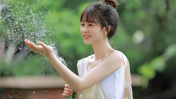 一名成年女性站在绿意盎然的户外环境中，微笑着伸手接住飞溅的水花。
