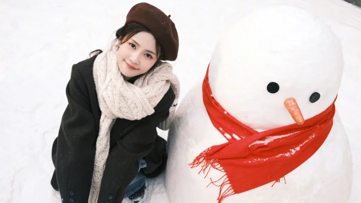 一名戴着棕色帽子和米色围巾的女子坐在雪地里的雪人旁边。