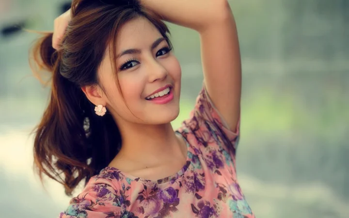 ぼかした背景の前で、花柄のトップスを着た成人女性が片腕を上げて微笑んでいる。