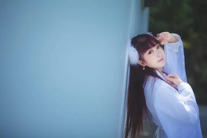 긴 머리와 앞머리를 한 여성이 흰색 옷을 입고 창가에 서서 카메라를 바라보고 있다.