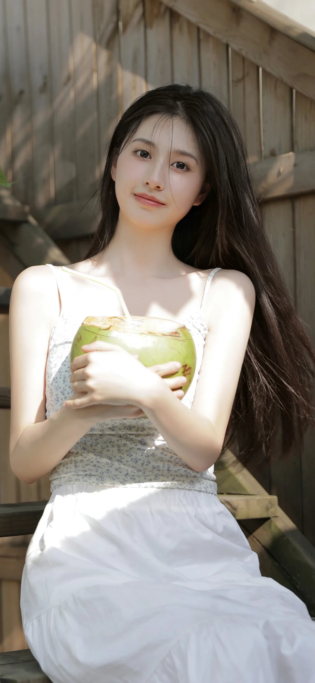一名长发女性穿着白色连衣裙坐在木墙旁的阳光下。