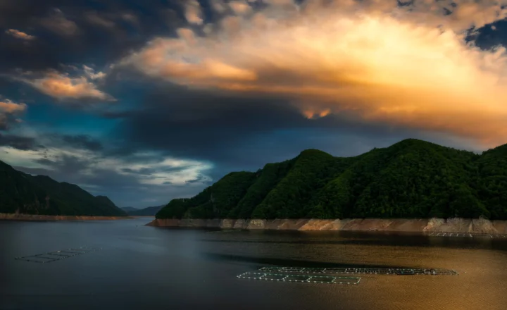 深绿色群山环绕着平静的湖面，戏剧性的云层在夕阳下泛着金色光芒，水面上可见养殖网箱。