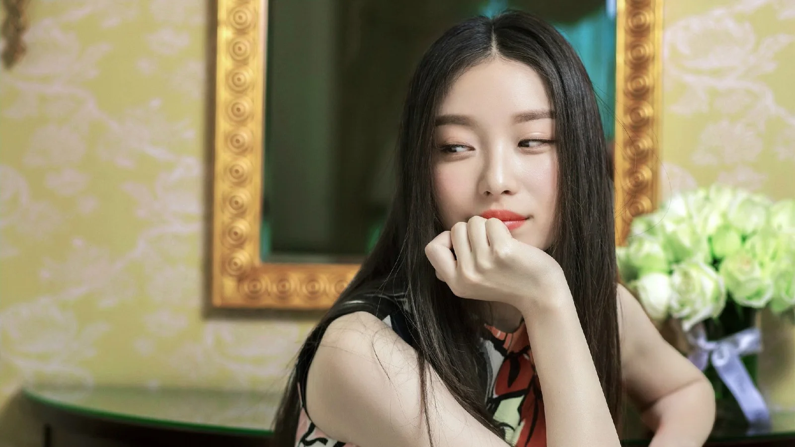 긴 머리의 여성이 실내에서 손으로 턱을 괴고 있으며 뒤에는 거울과 꽃다발이 보입니다.