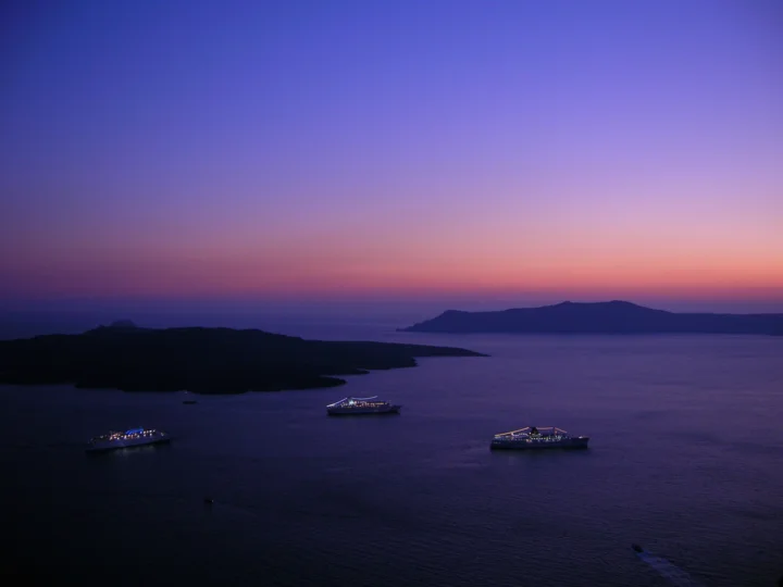 暮色中的海湾映着紫蓝与粉色天空，远处是山峦剪影，海面上停泊着几艘亮灯的船。
