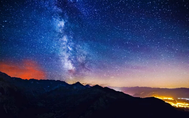 璀璨的银河横跨繁星密布的夜空，映照着下方深色的山脉轮廓，远处山谷闪烁着温暖灯光。