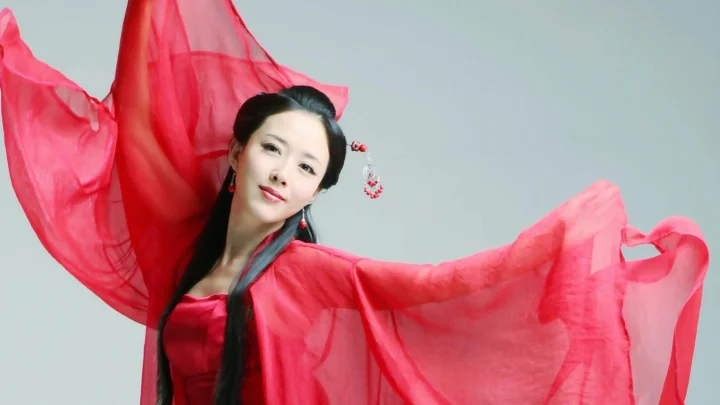 붉은 전통 의상을 입은 성인 여성이 연한 회색 배경 앞에서 흘러내리는 소매를 펼치고 있다.