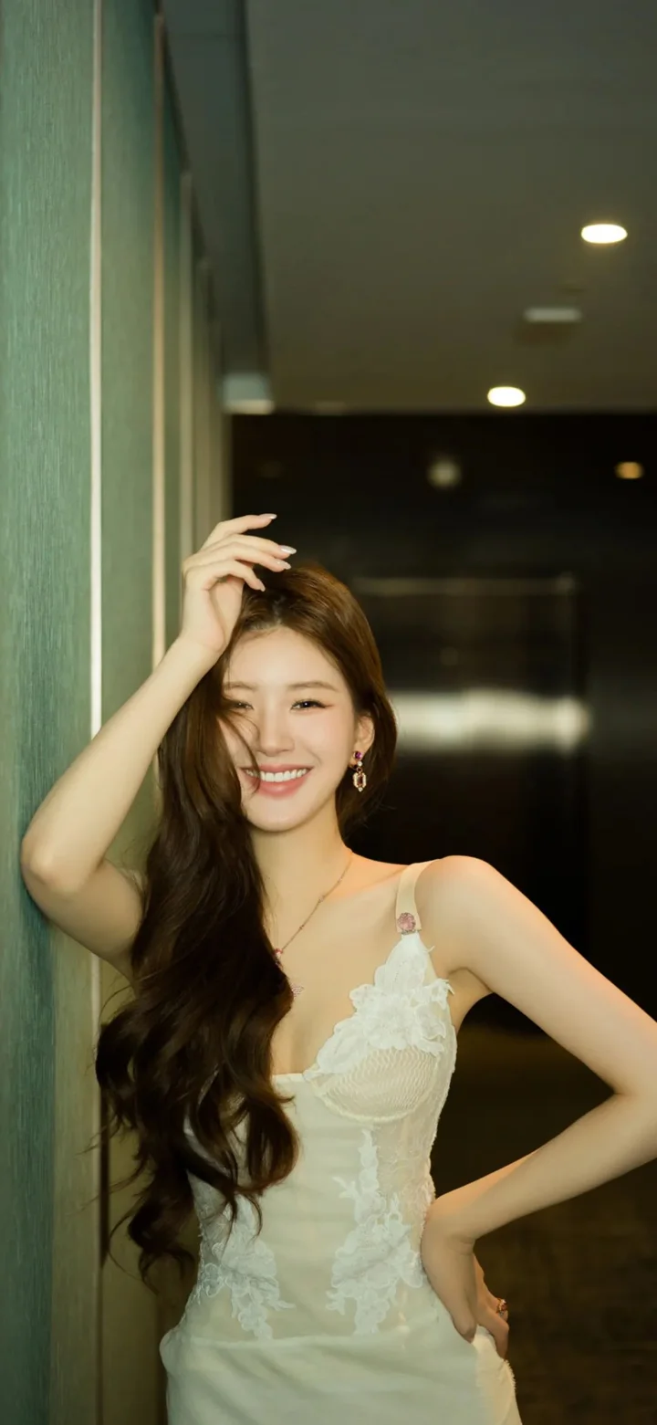 一名长发女性身穿白色连衣裙站在室内走廊中微笑面对镜头。