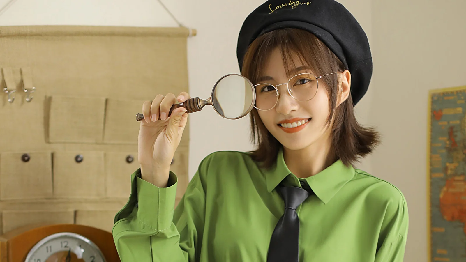 검은 베레모와 둥근 안경을 쓴 여성이 초록색 셔츠를 입고 돋보기를 들고 카메라를 향해 미소 짓고 있다.