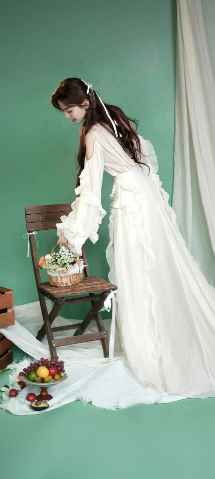 白いロングドレスを着た女性が、緑色の背景のスタジオで椅子の上の花かごのそばに身をかがめています。