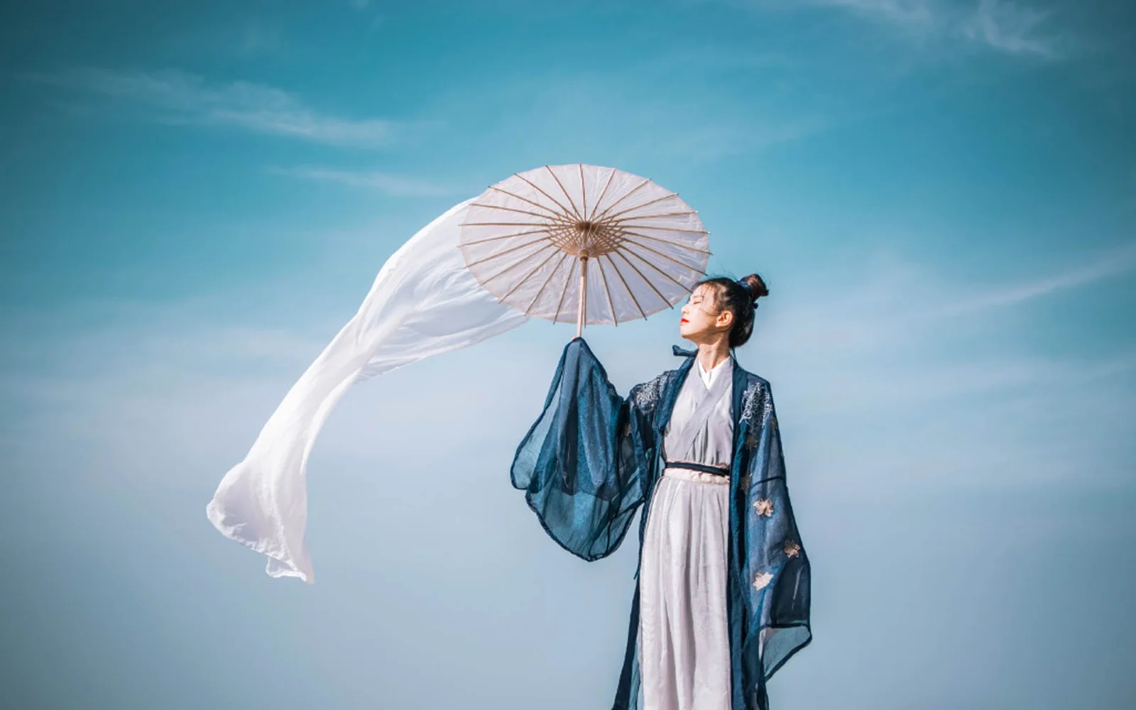 一名身穿蓝白色传统服饰的女子撑着纸伞，伞边白色布料在蓝天前飘动。