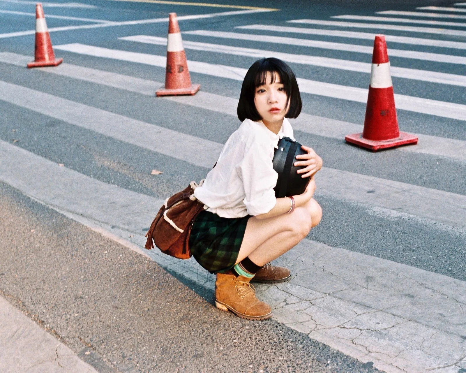 短い髪の女性が黒い物を抱えて横断歩道のそばにしゃがみ、周囲にはカラーコーンが置かれている。