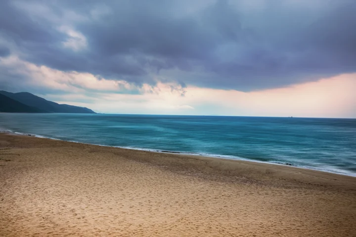 沙滩、湛蓝海水与远处山脉延伸在阴云笼罩的海岸线上。