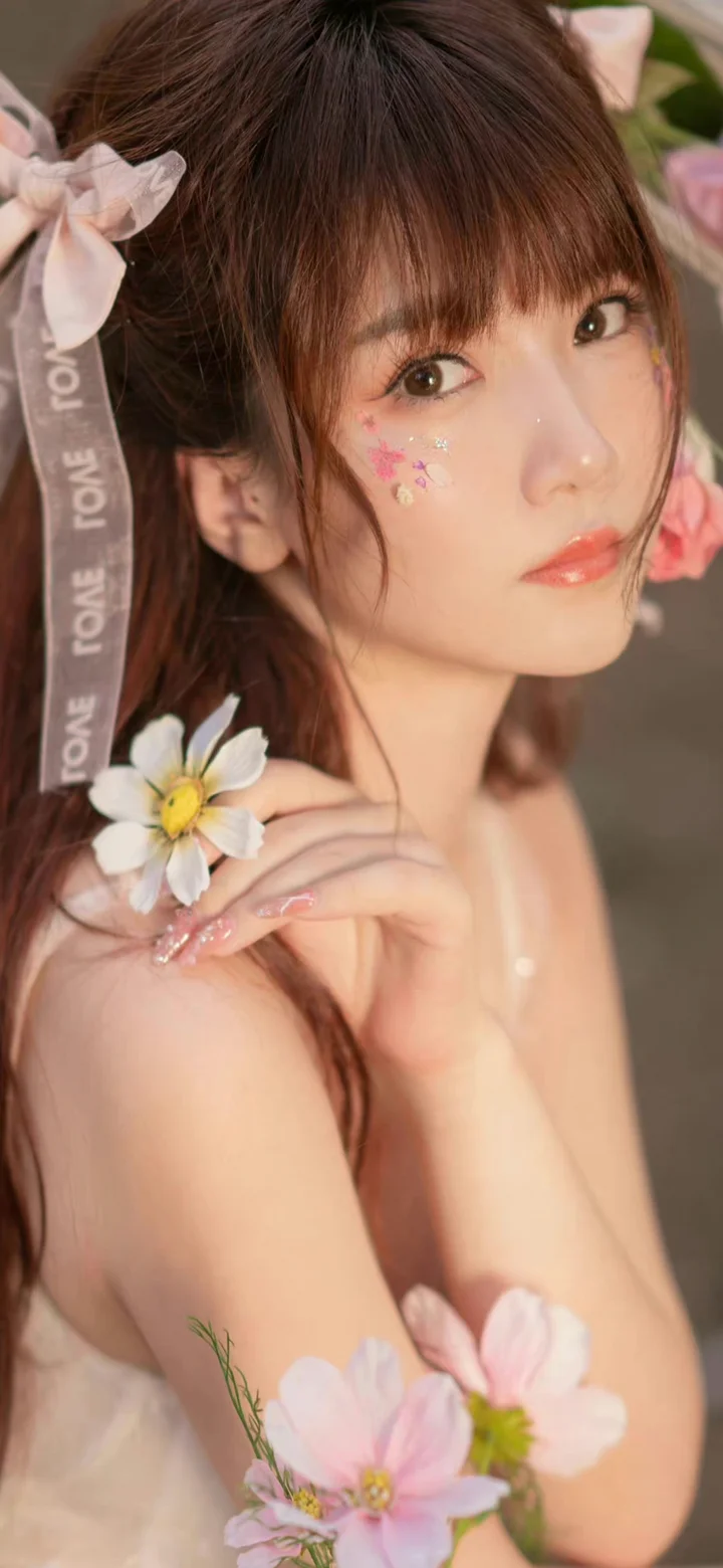 一位留着棕色长发的女性手持白色小花，脸颊点缀花朵贴纸，画面柔和清新。