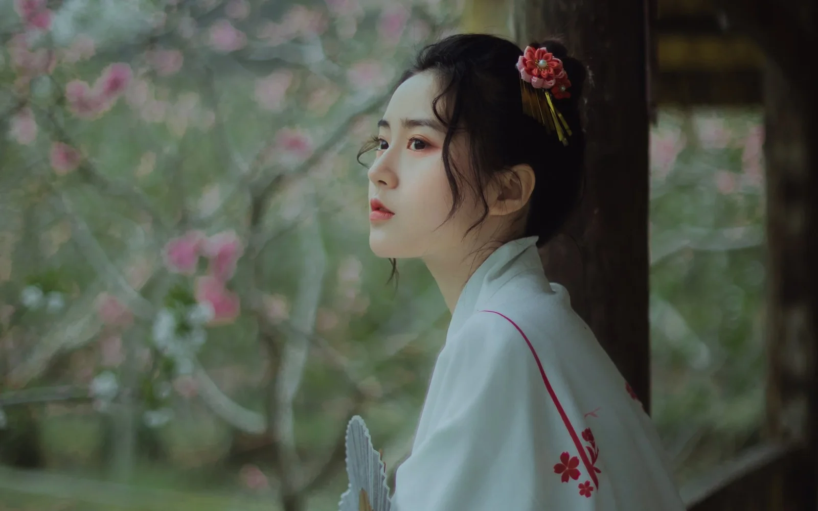밝은 색상의 전통 의상을 입은 여성이 꽃나무와 나무 창문 옆에서 뒤돌아보고 있다.