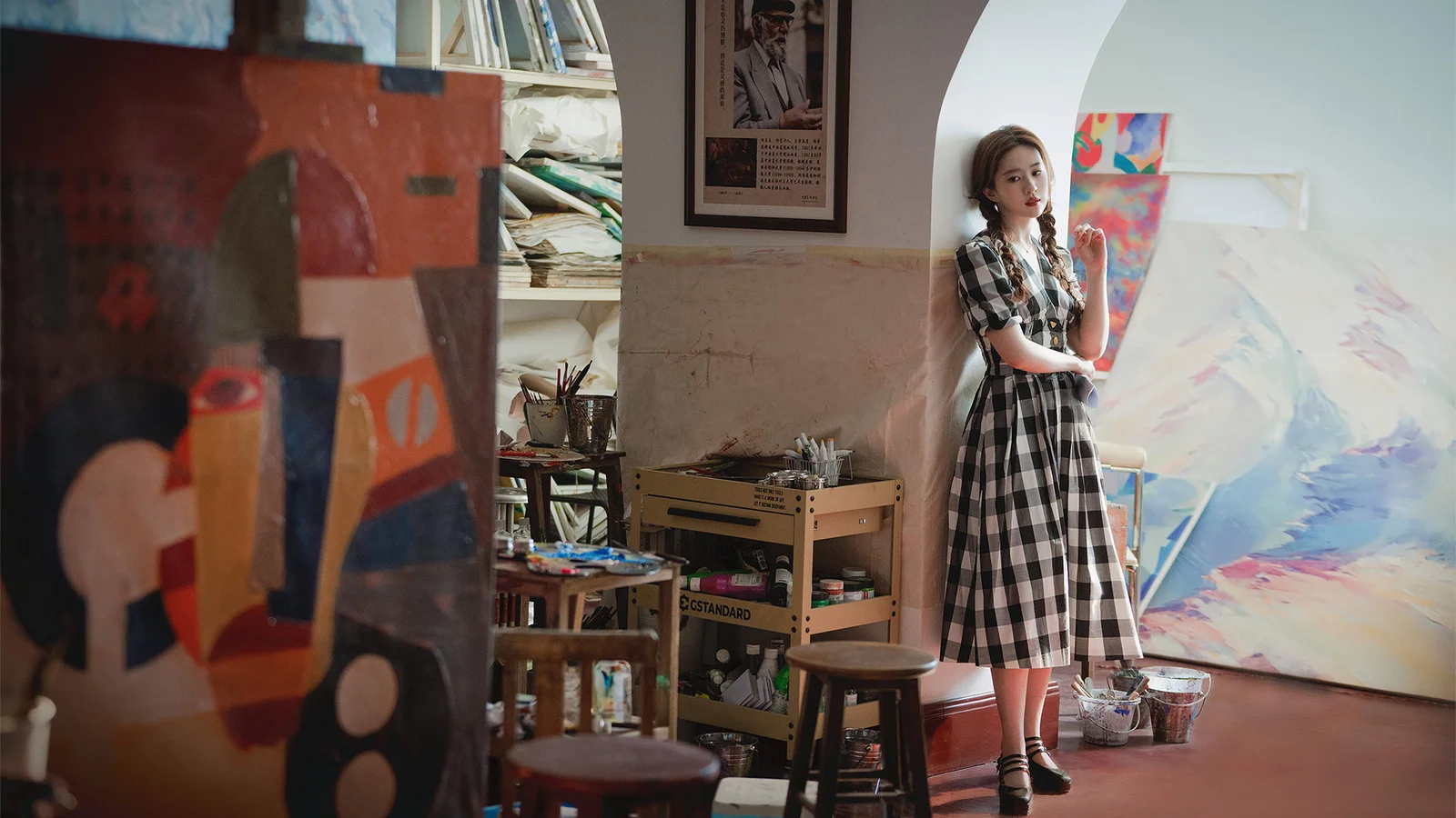 明るいアトリエで、絵画やイーゼル、画材に囲まれて女性が立っています。