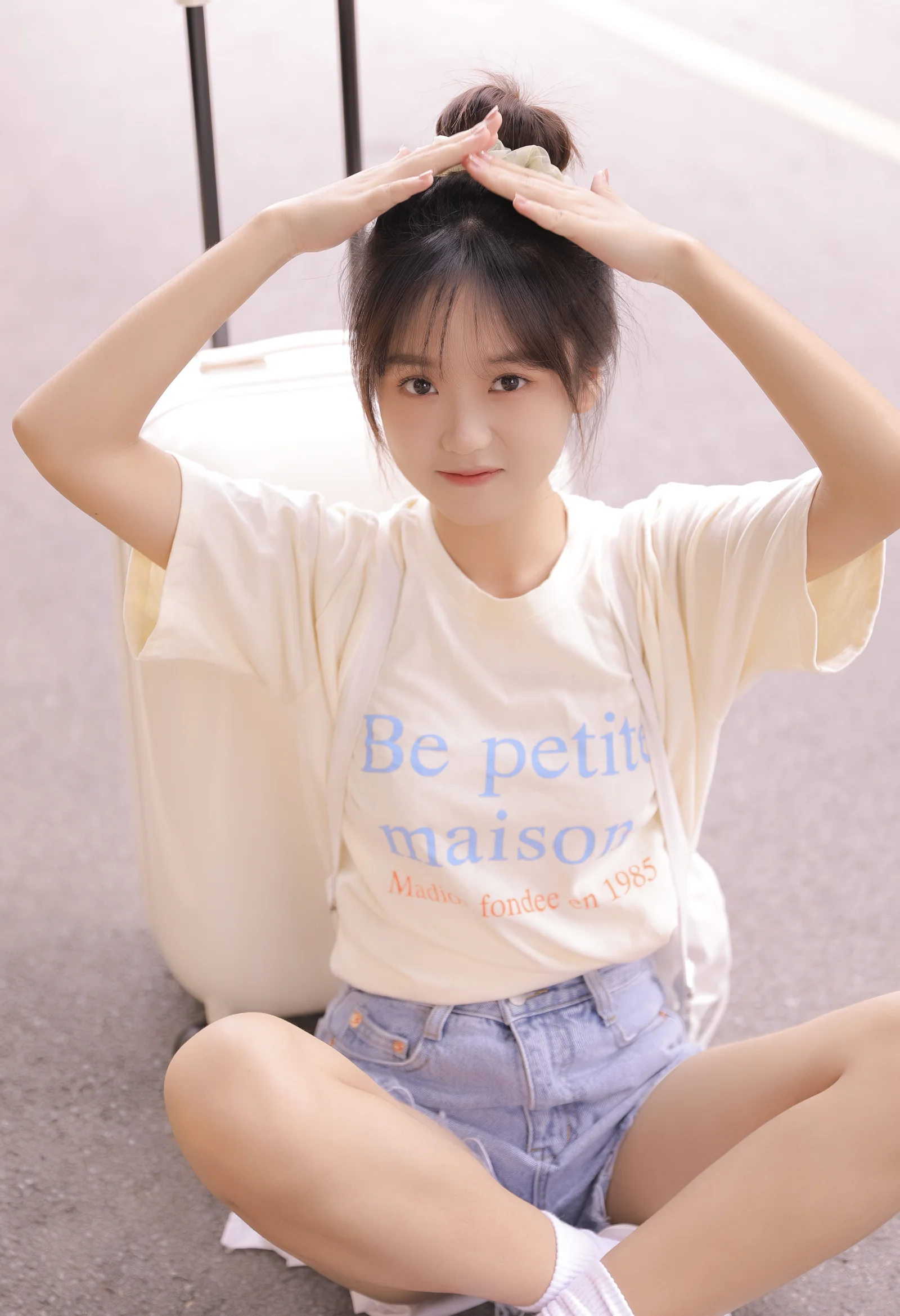 높은 포니테일을 한 젊은 여성이 실내에 앉아 두 손을 머리 위로 올리고 귀여운 포즈를 취하고 있다.