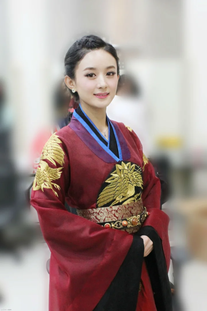 一名成年女性身穿带金色纹样的红色传统服饰，站在虚化背景前。