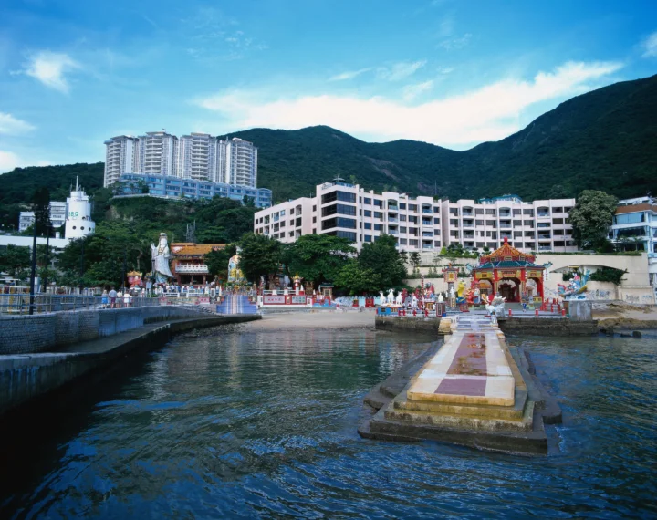 一座滨水寺庙坐落在高楼与青山之间，前方水面倒映着建筑和天空。