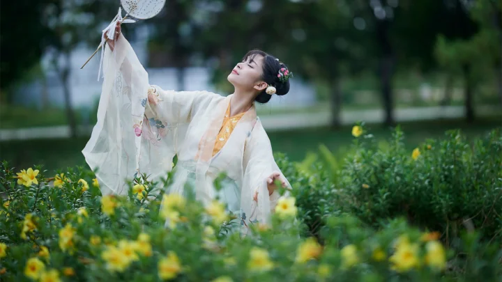 一名身穿传统服饰的女子站在户外花丛中，手持装饰物并仰头望向上方。