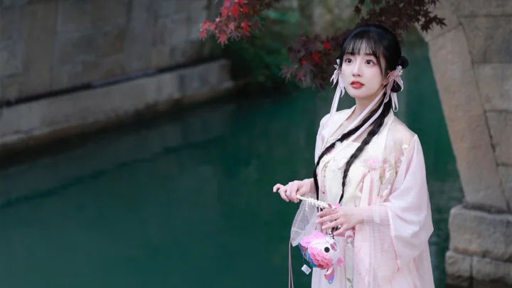 一名身穿浅粉色汉服的女子站在绿色水面与石墙旁。