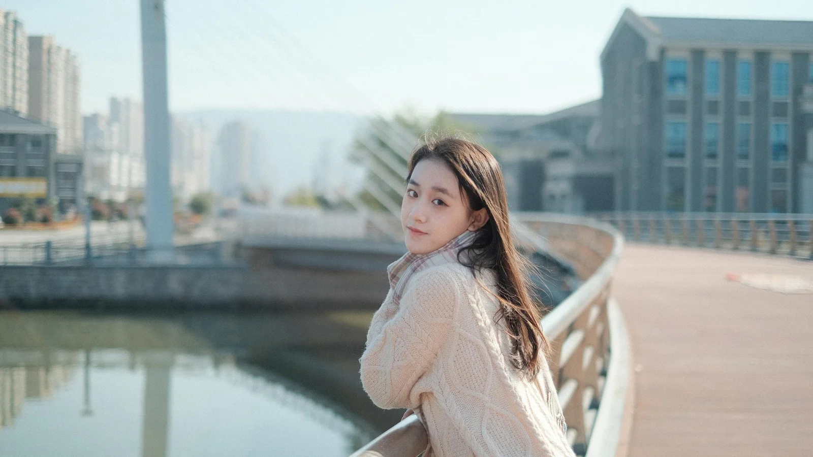 一名长发女性穿着浅色针织衫站在城市桥边回眸，背景是水面、建筑和步道。