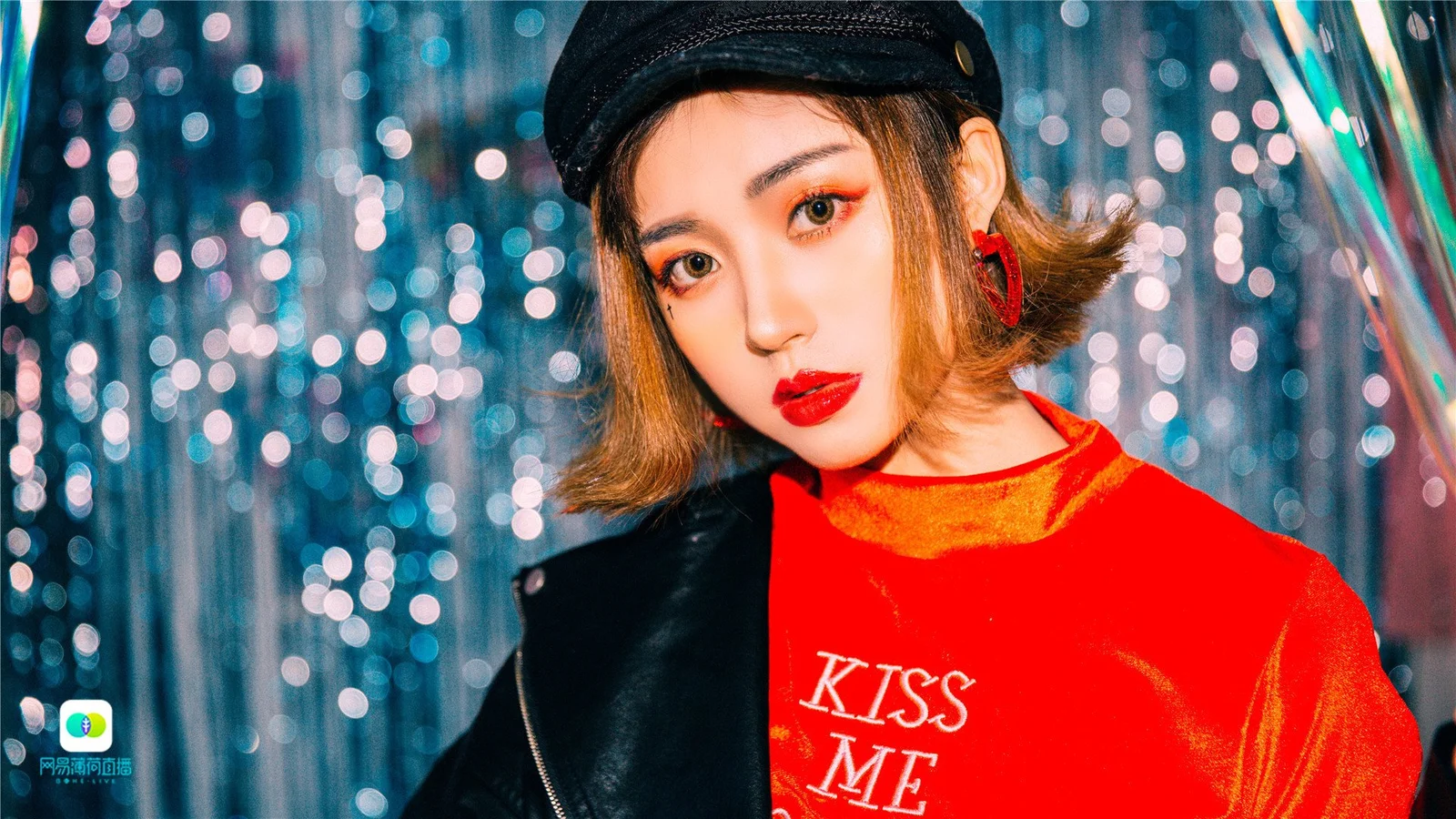 검은 모자와 빨간 셔츠를 착용한 성인 여성이 반짝이는 배경 앞에 서 있습니다.