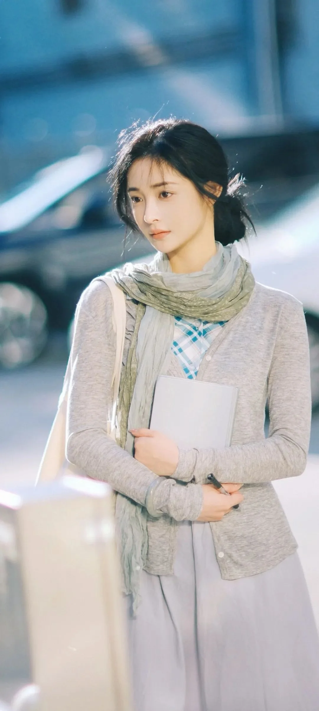 스카프를 두른 여성이 흰색 노트를 들고 흐릿한 실내 배경 앞에 서 있다.