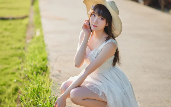 一名戴着宽檐帽、穿白色连衣裙的女子坐在草地旁的步道上。