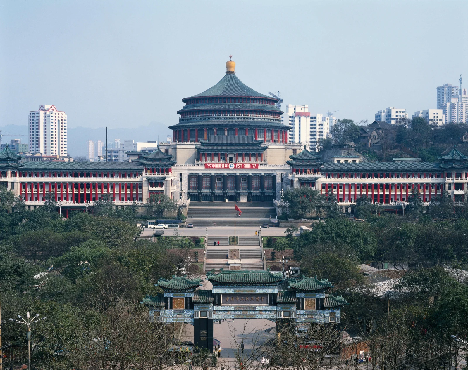 一座大型中式传统建筑坐落在绿树之间，背景是城市高层建筑和晴朗天空。