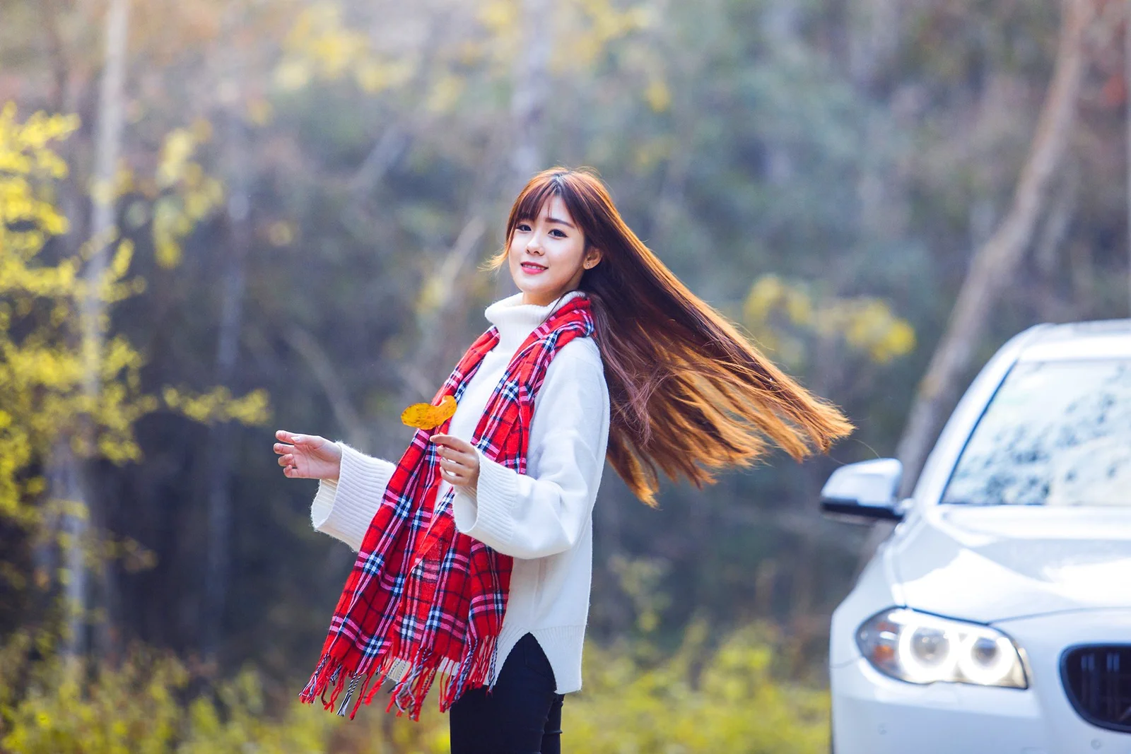 一名长发女子站在树林边，系着红色格纹围巾，旁边停着一辆白色汽车。