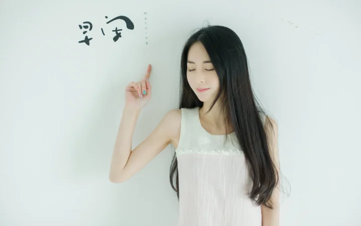 一名身穿白色无袖连衣裙的女子闭着眼睛站在浅色背景前，并抬手指向上方。